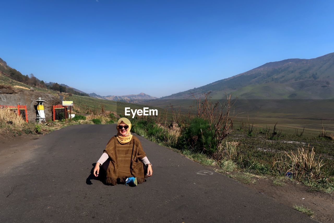 WOMAN SITTING ON ROAD AGAINST MOUNTAIN