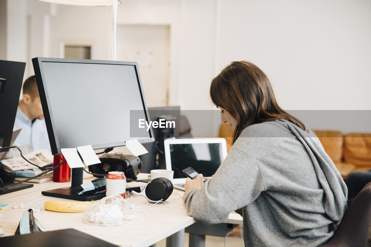 Side view of female programmer using mobile phone on desk while sitting in office