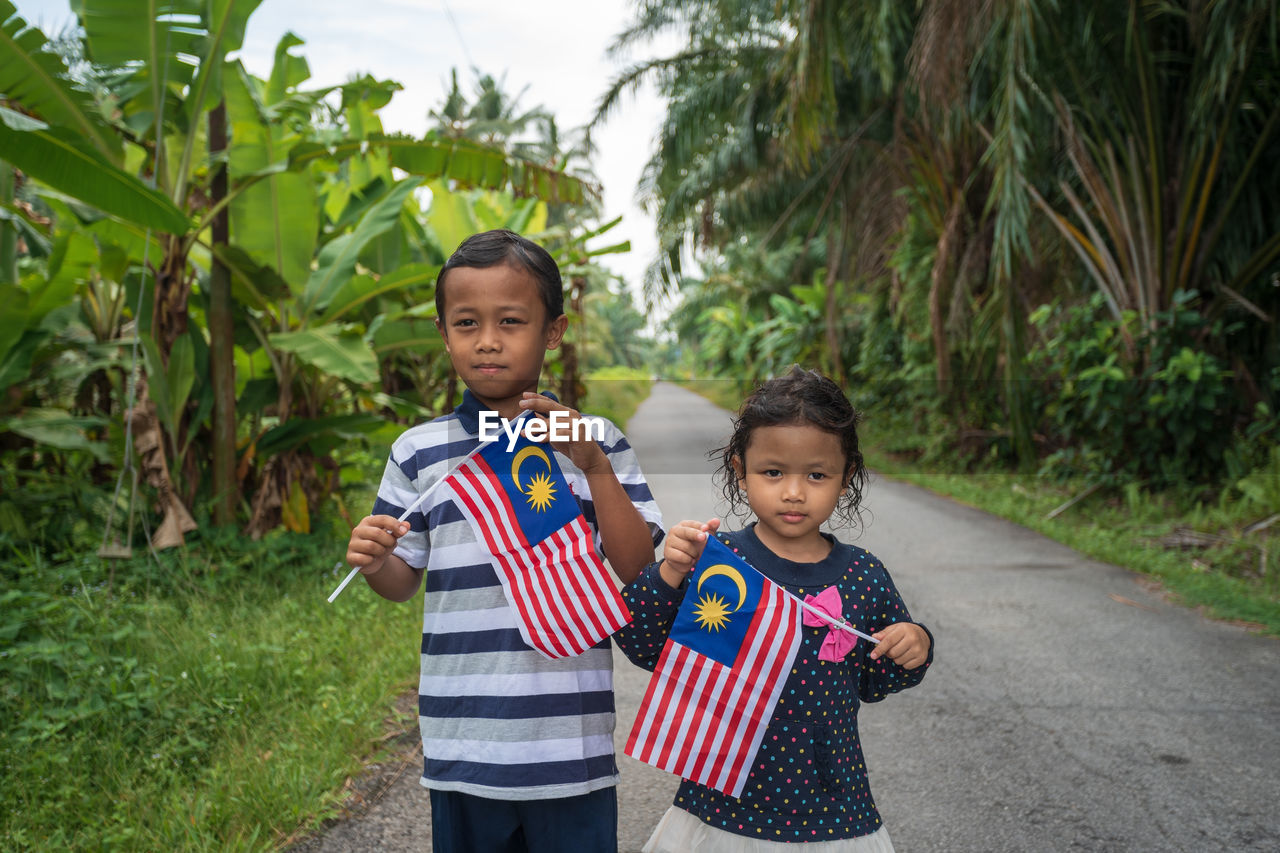 Siblings holding malaysian flag while standing on road amidst plants
