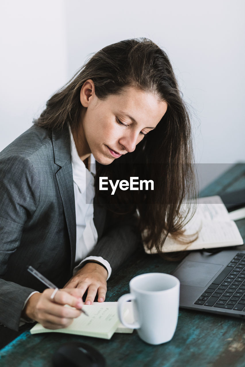 Side view of focused young female entrepreneur in formal wear sitting at table with laptop and cup of coffee and writing information in notebook while planning working day at workplace at home