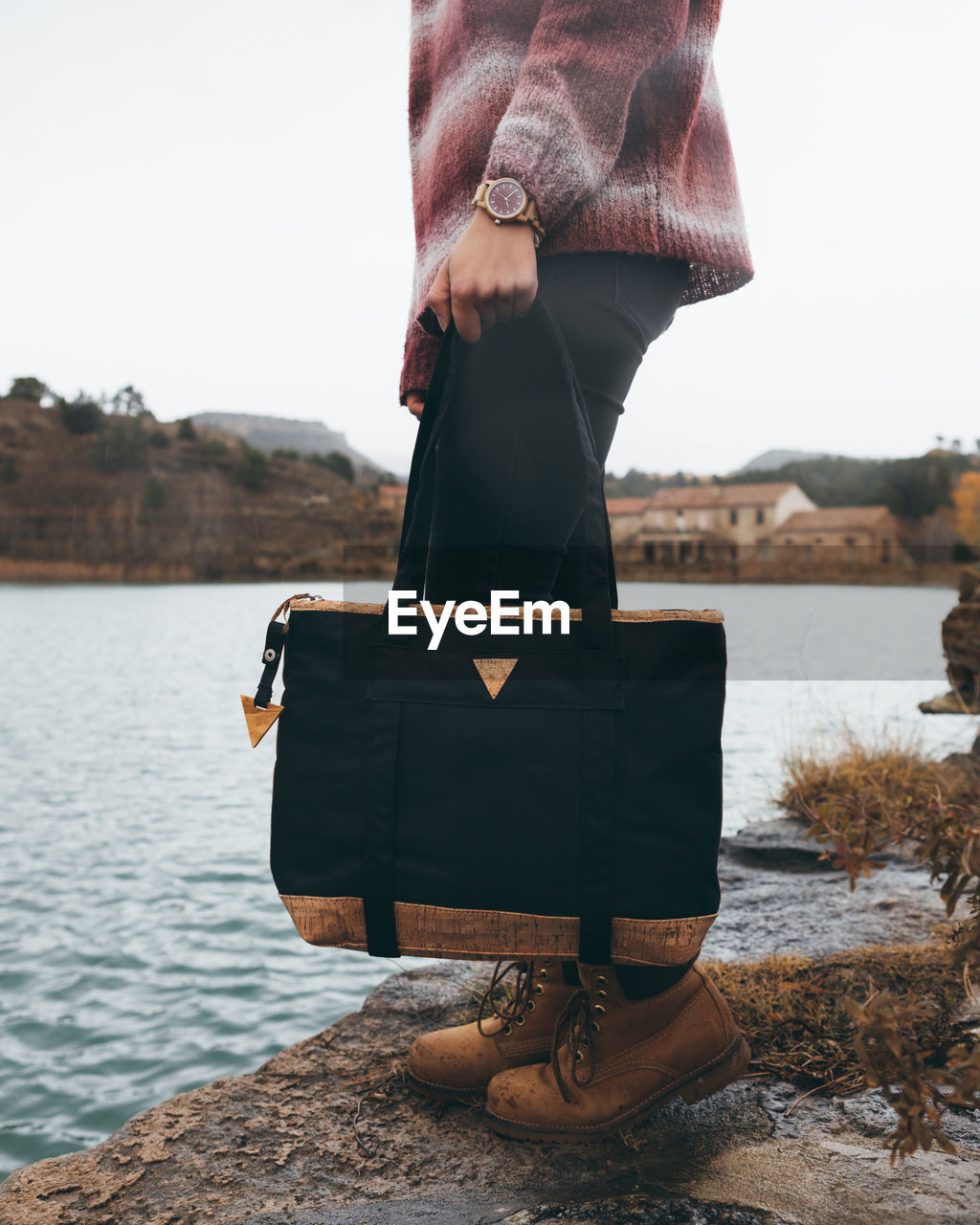 Side view of crop stylish female in casual outfit with trendy black handbag standing on riverbank in town in cloudy cold autumn day