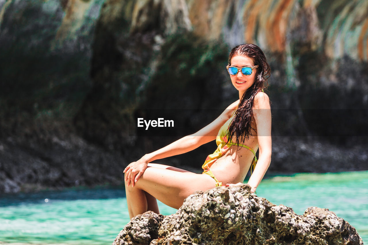 Seductive woman wearing swimsuit sitting on rock by sea during summer