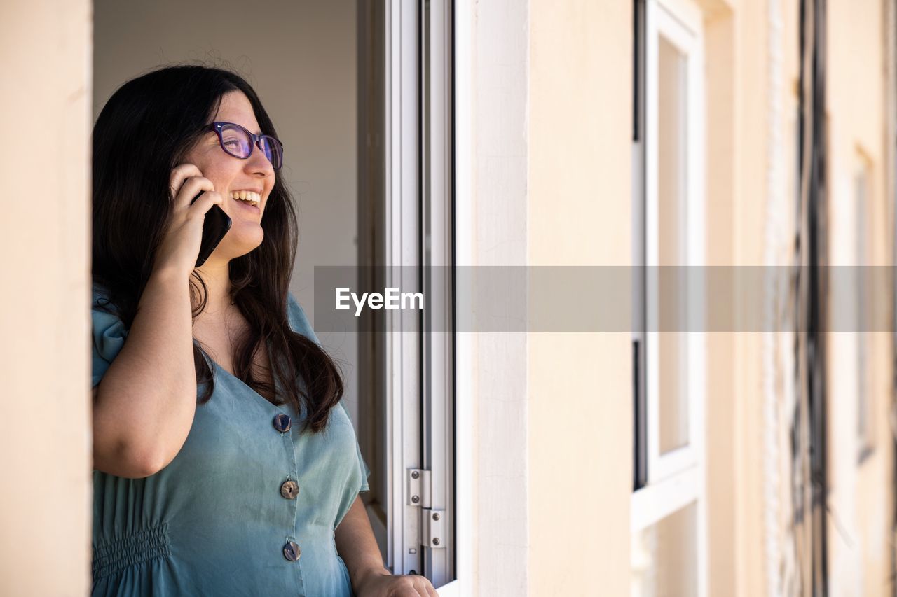 Brunette spanish girl dressed on a business serius look talking on the phone at the window from her apartment during the afternoon in palma de mallorca, spain during coronavirus confinement