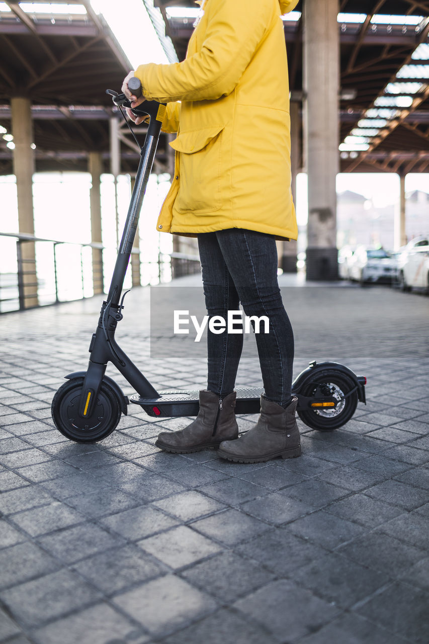 Young woman in yellow coat prepares and uses her electric skateboard.