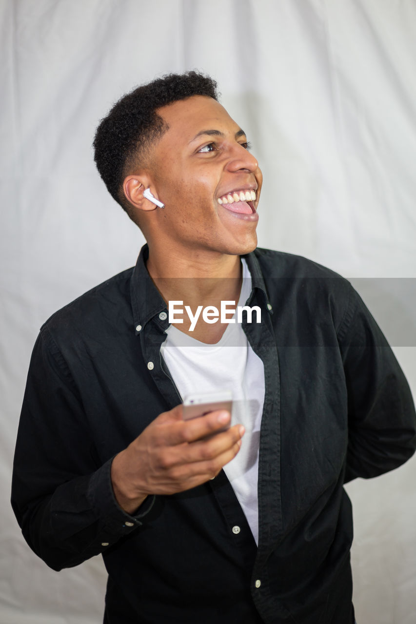 SMILING YOUNG MAN LOOKING AWAY WHILE STANDING AGAINST WALL