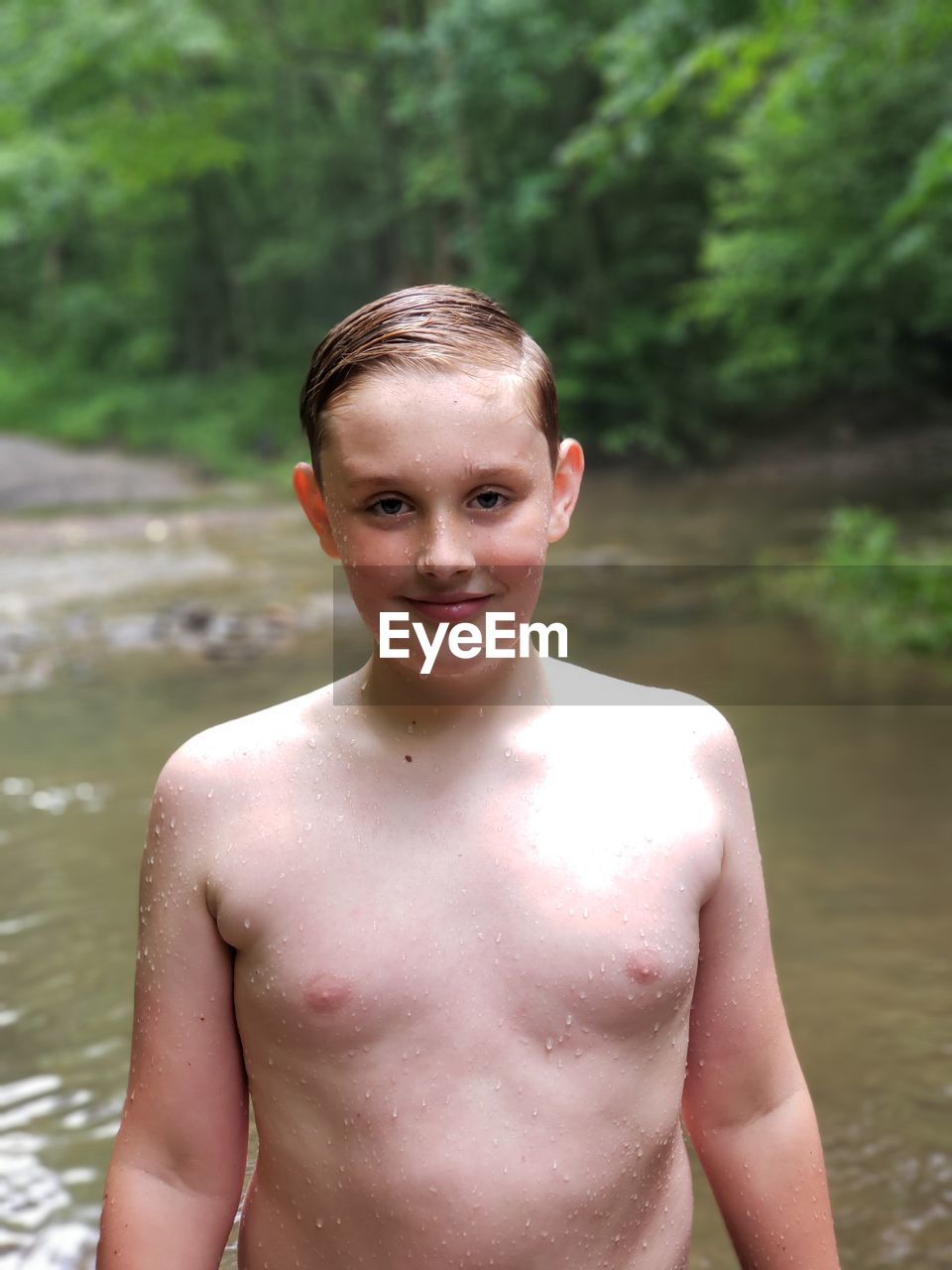 Portrait of smiling shirtless boy standing against stream in forest