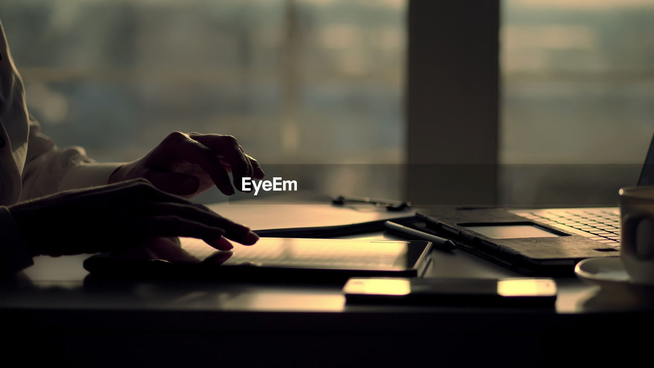 Dark silhouette of female hands. she is typing something in tablet, next is a cup, laptop on table.