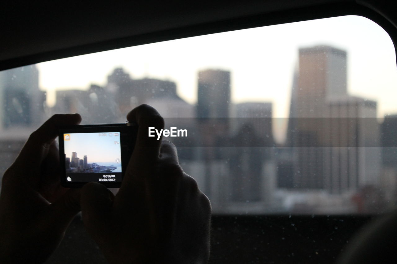 Cropped image of hand photographing cityscape seen through window
