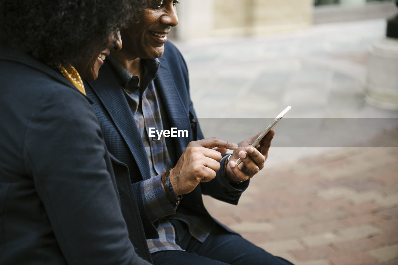 Happy woman looking at man using mobile phone in city