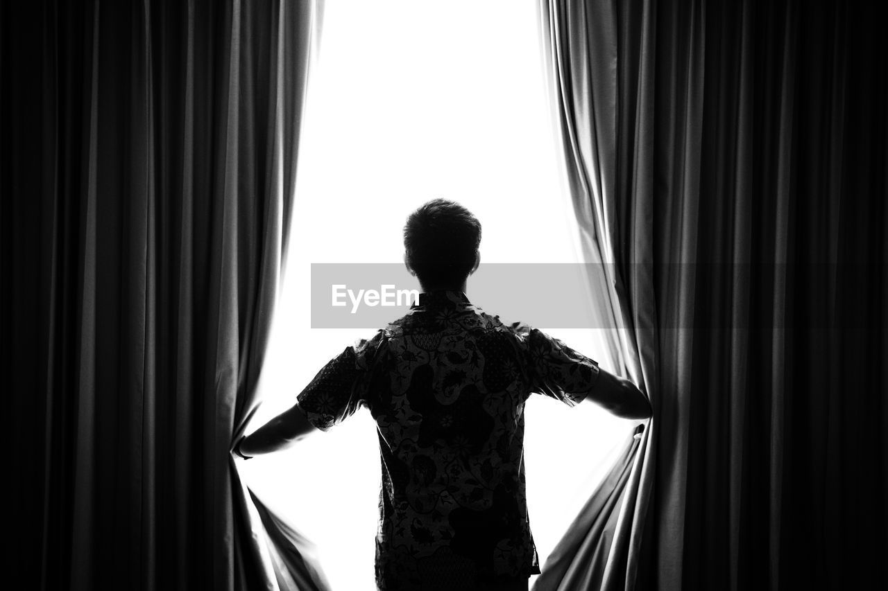 Rear view of man opening curtains on window at home