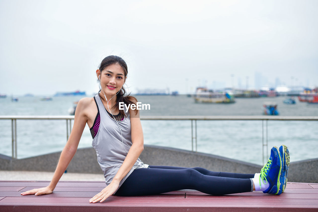 Portrait of young woman sitting on bench against sea and sky