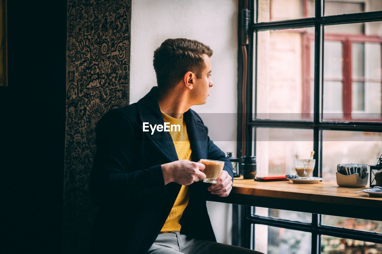 Young man holding coffee looking through window while sitting in cafe