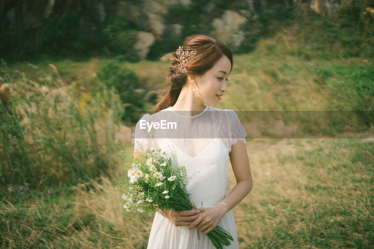 Bride holding flowers looking away while standing on grassy field