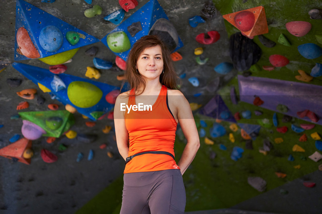Charming strong woman in sportswear holding hands behind back while standing against climbing wall and looking at camera