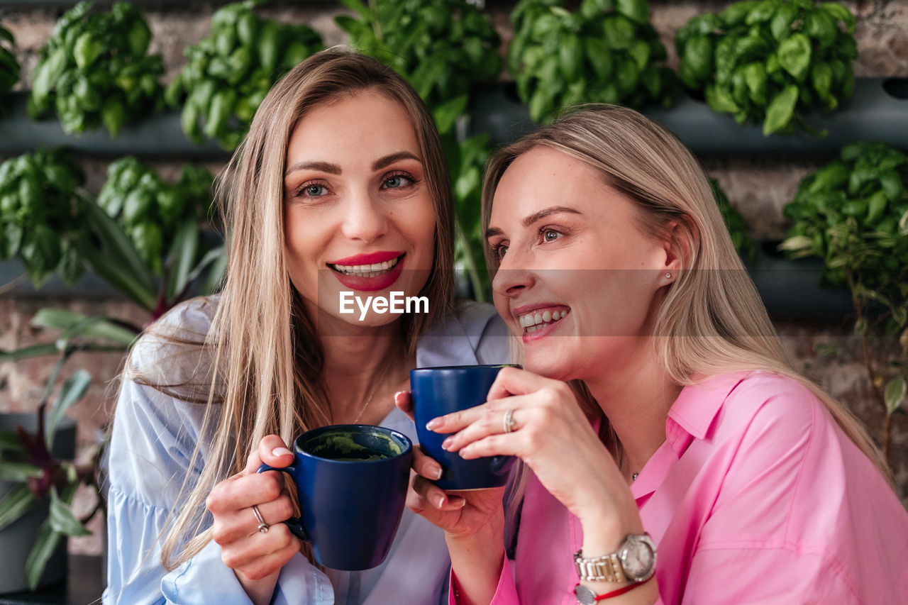 women, smiling, adult, happiness, two people, togetherness, drink, portrait, emotion, food and drink, young adult, cup, female, friendship, blond hair, mug, lifestyles, coffee, cheerful, refreshment, enjoyment, drinking, bonding, positive emotion, leisure activity, holding, communication, headshot, coffee cup, person, relaxation, casual clothing, love, hot drink, portable information device, long hair, smartphone, teeth, looking at camera, wireless technology, outdoors, smile, fun, nature, clothing, food, hairstyle, technology, sitting, summer, human face, camera, ceremony, front view, talking