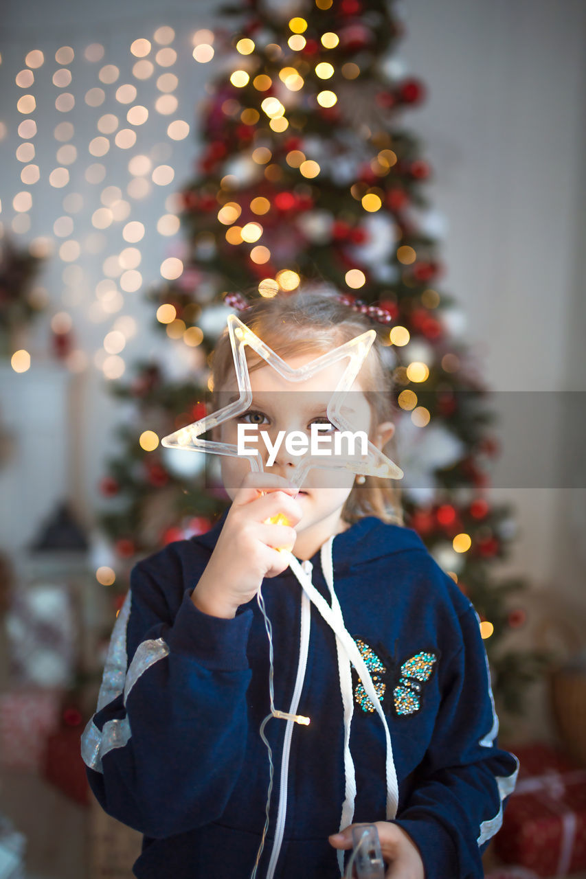 Portrait of cute girl holding star shape standing against christmas tree at home