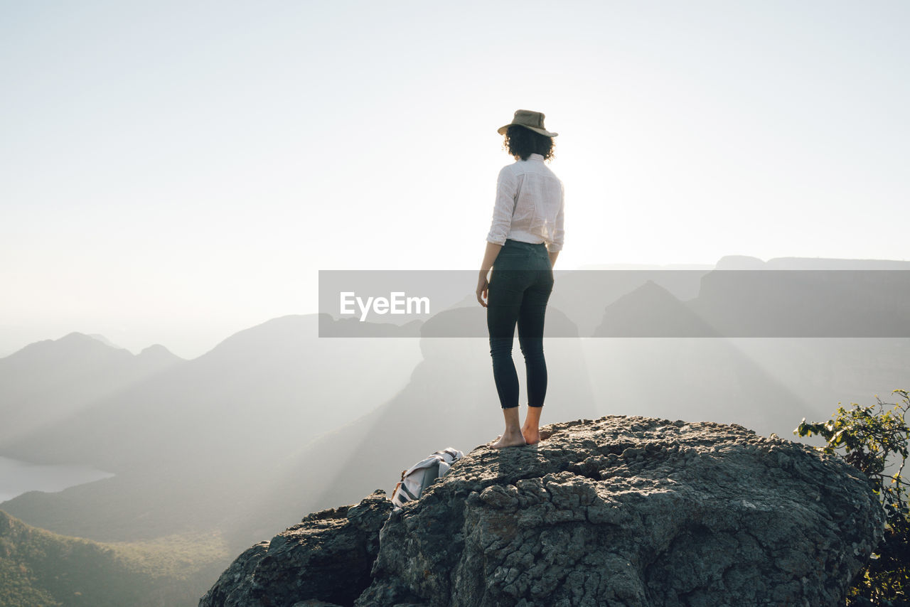 Rear view of woman looking at view while standing on rock against sky during sunny day