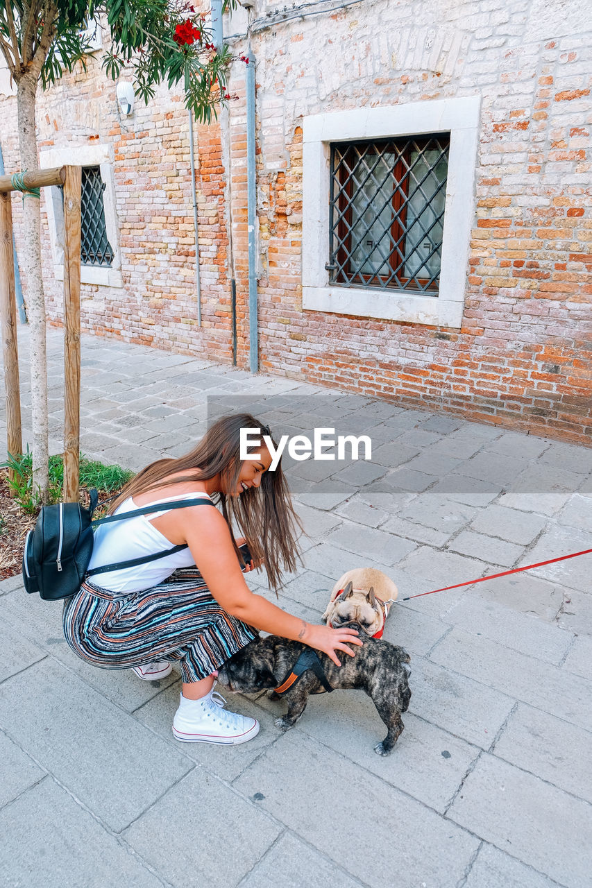 Side view of young woman with backpack petting dogs on footpath