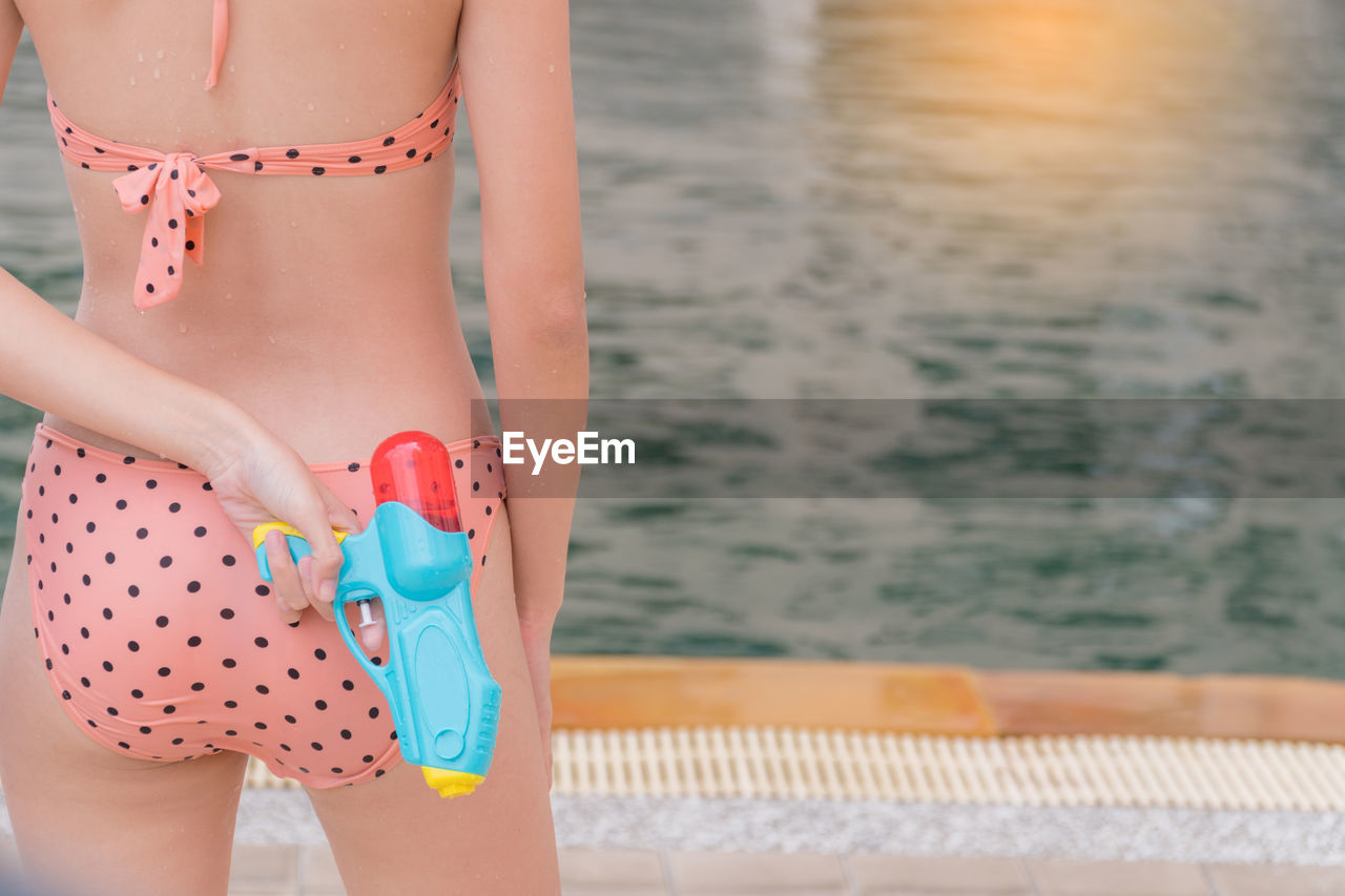 Rear view midsection of woman wearing bikini while holding squirt gun on poolside