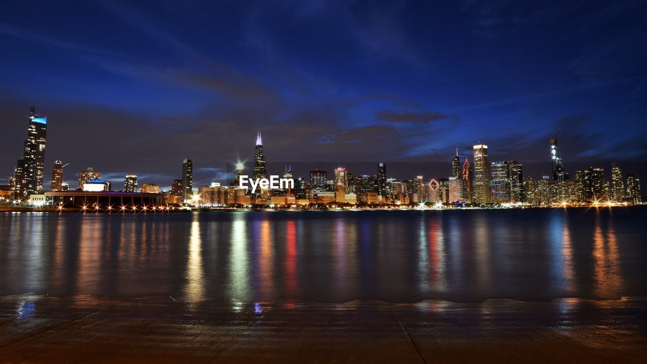 Illuminated buildings by river against sky at night