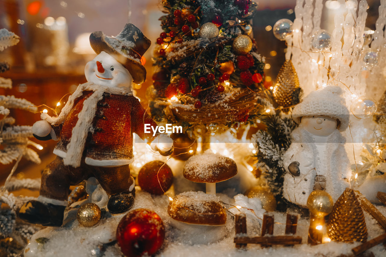 Funny winter toy snowman and snow covered christmas tree with golden lights on a festive storefront