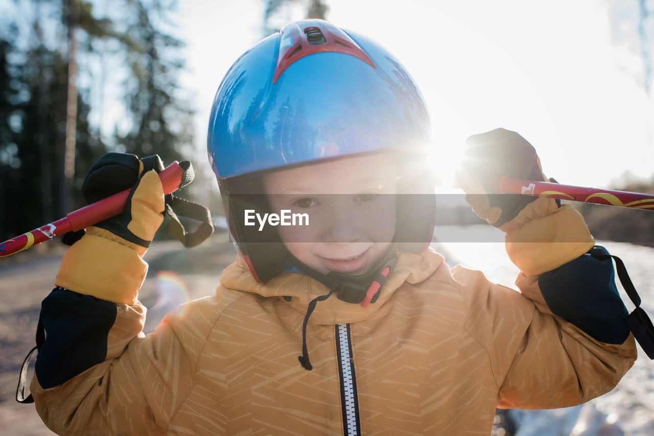 Close up of a young boy wearing a ski helmet holding poles at sunset