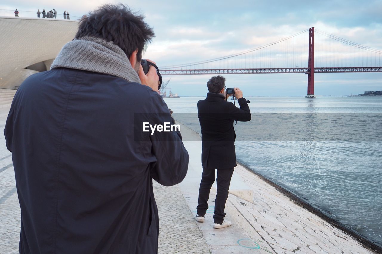 Rear view of people photographing suspension bridge while standing by river