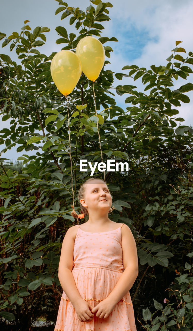 Girl in a pink dress stands on the street. yellow balloons are tied to her braided hair.