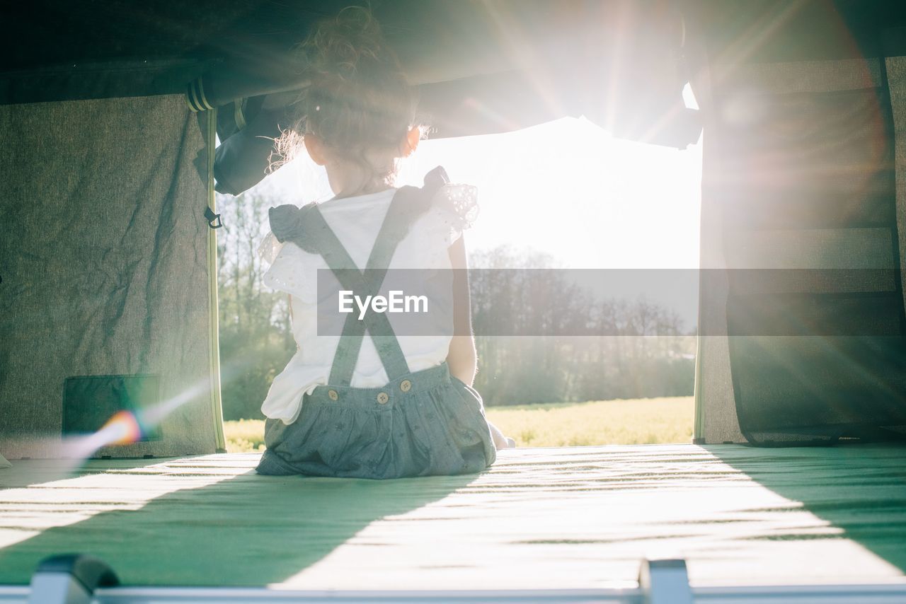 Young girl sat in a tent watching the sun set whilst camping outdoors