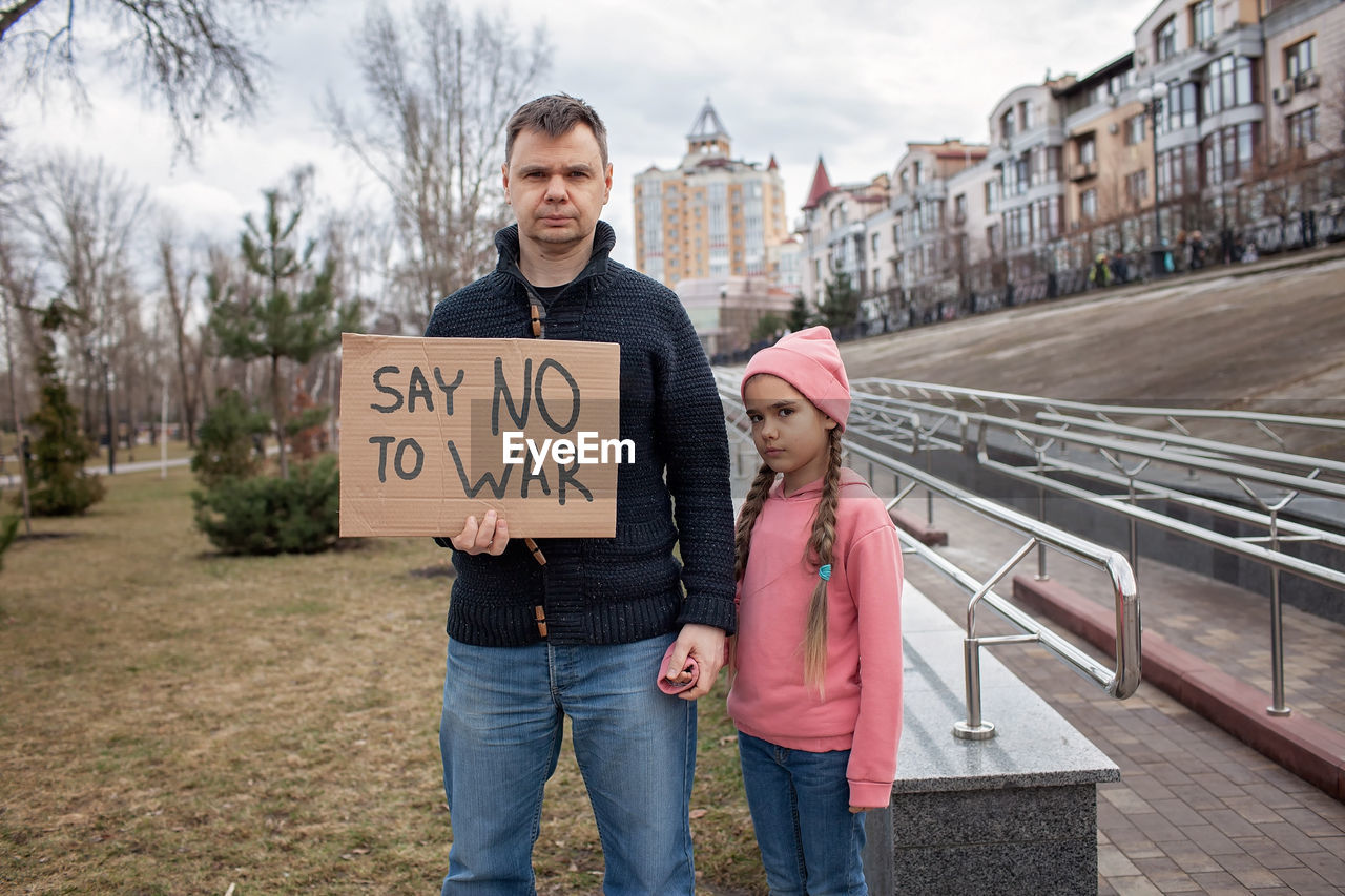 Middle aged father with daughter holding a poster with anti-war message over cityscape background