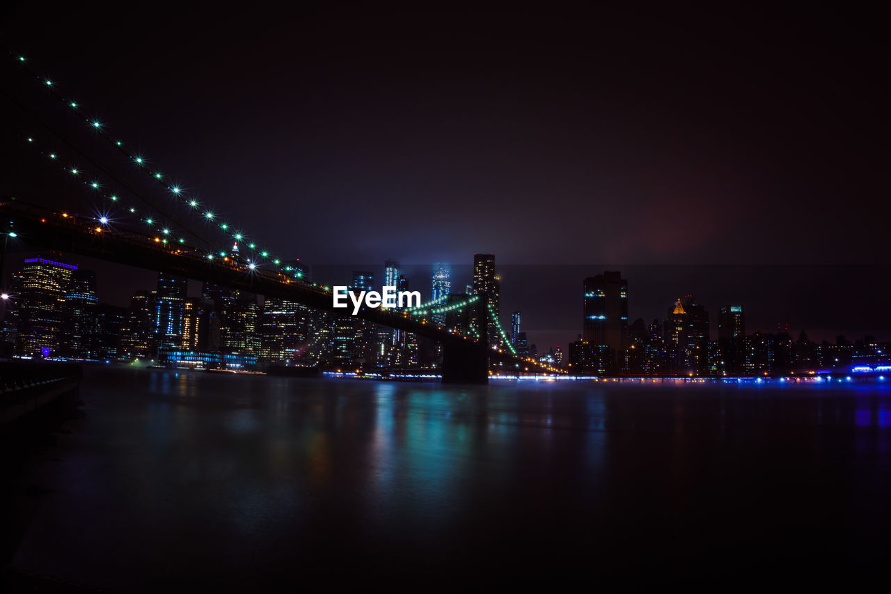 Illuminated modern buildings by river against sky at night