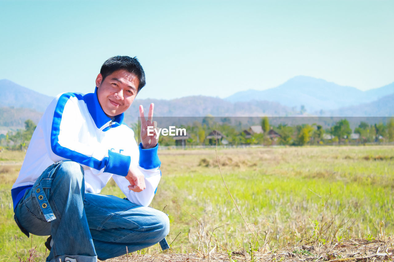 Portrait of smiling man showing peace sign while crouching in field