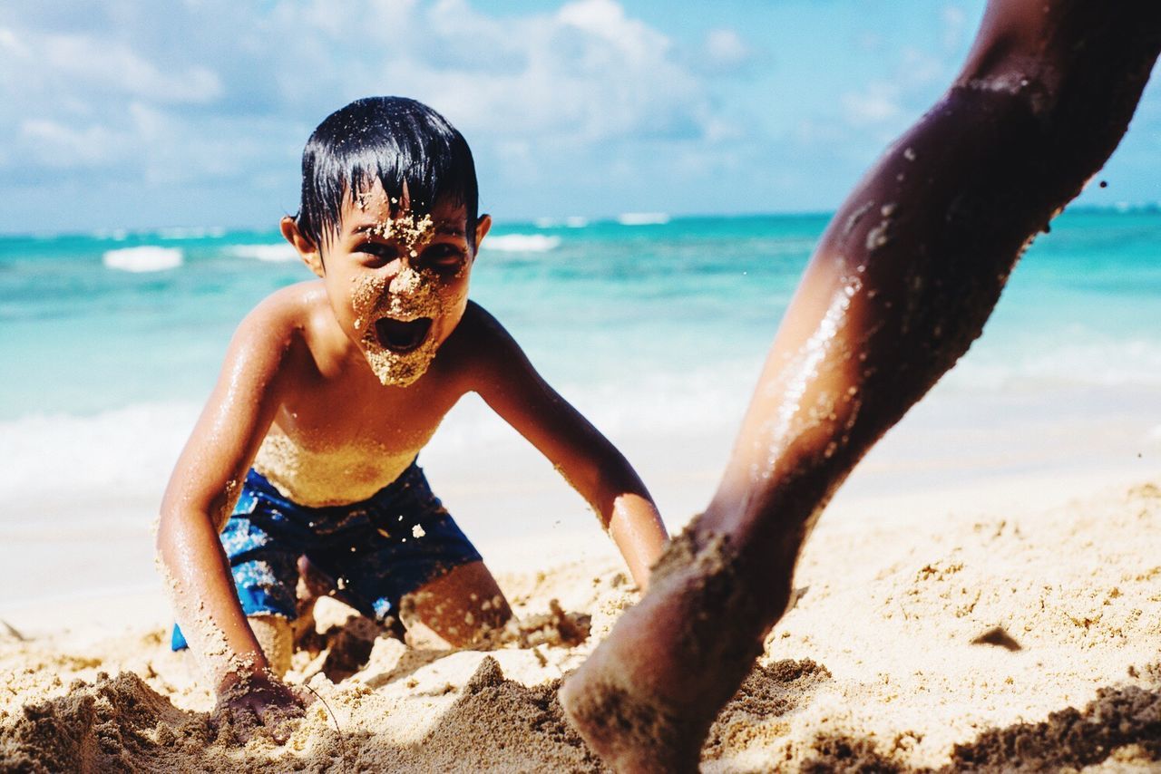 Boy with sand on face screaming while kneeling at beach