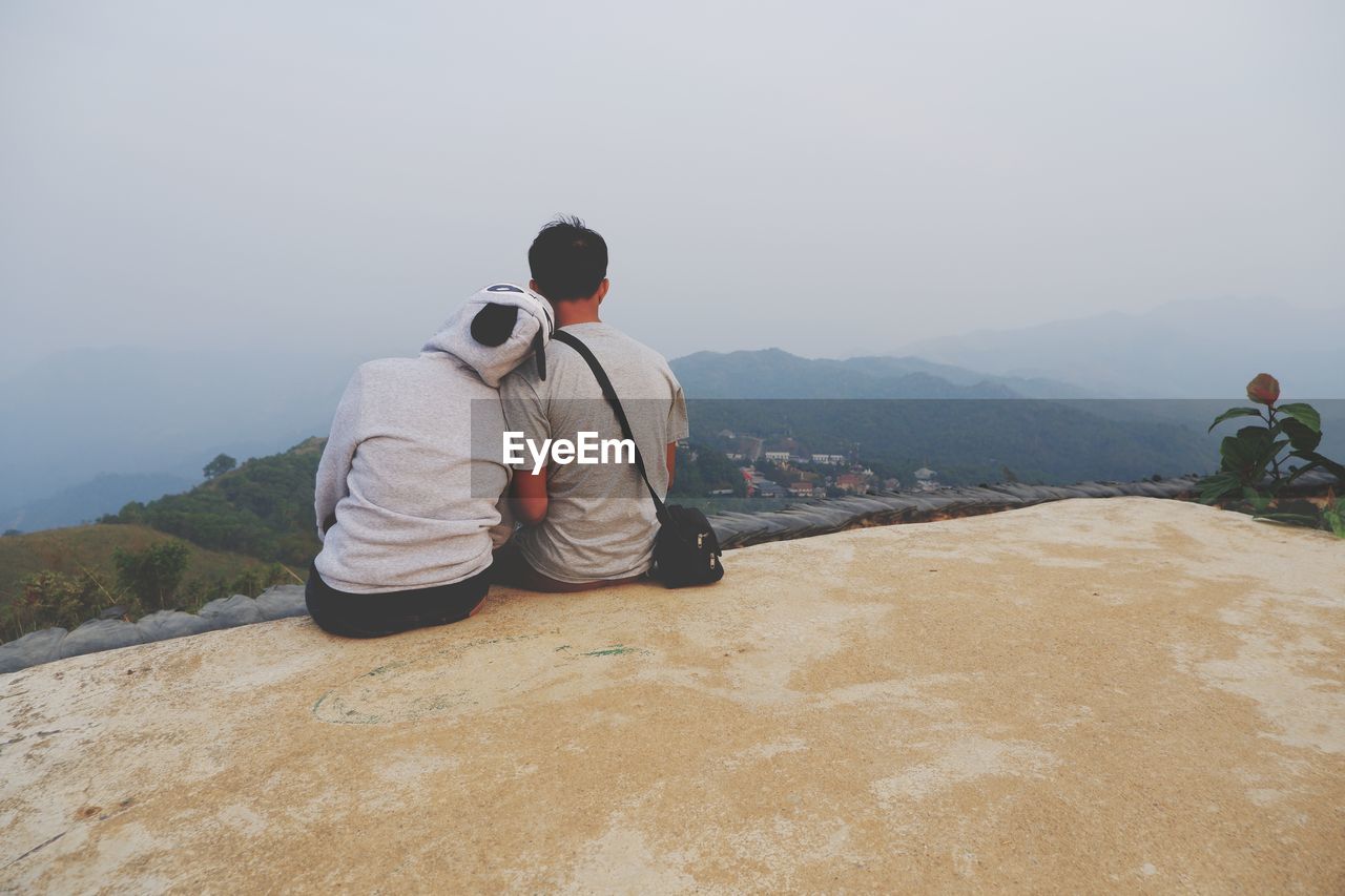 REAR VIEW OF MAN SITTING ON MOUNTAIN LOOKING AT VIEW