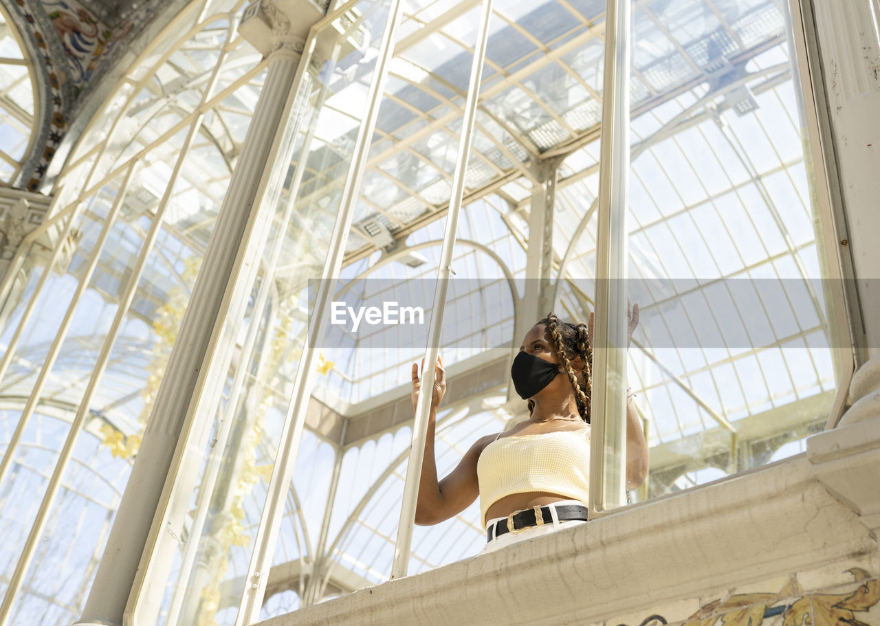 Trendy ethnic female with braids wearing protective mask standing in glass building lit by sunlight while looking through window