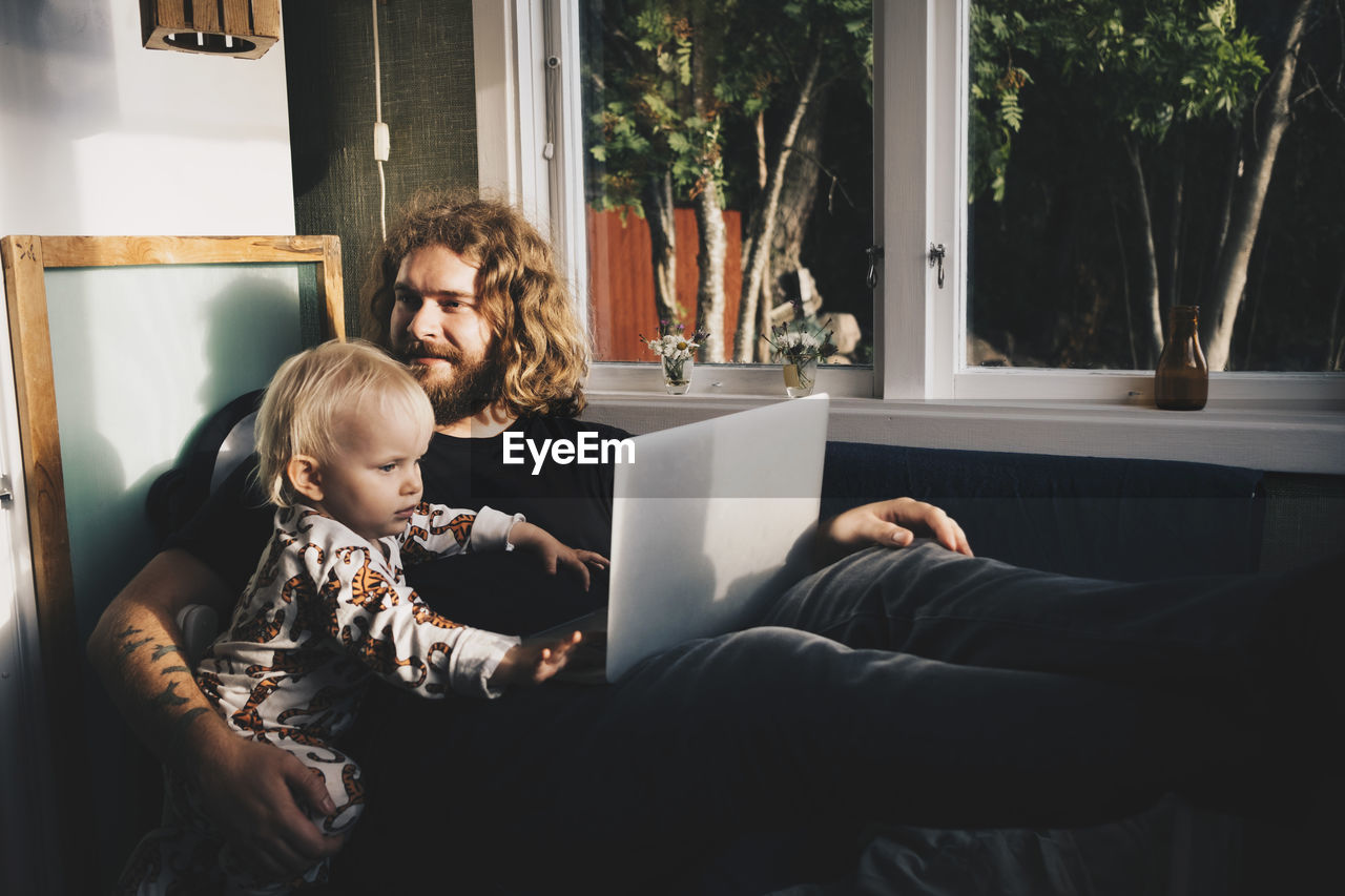 Father with girl holding laptop while sitting by window at home