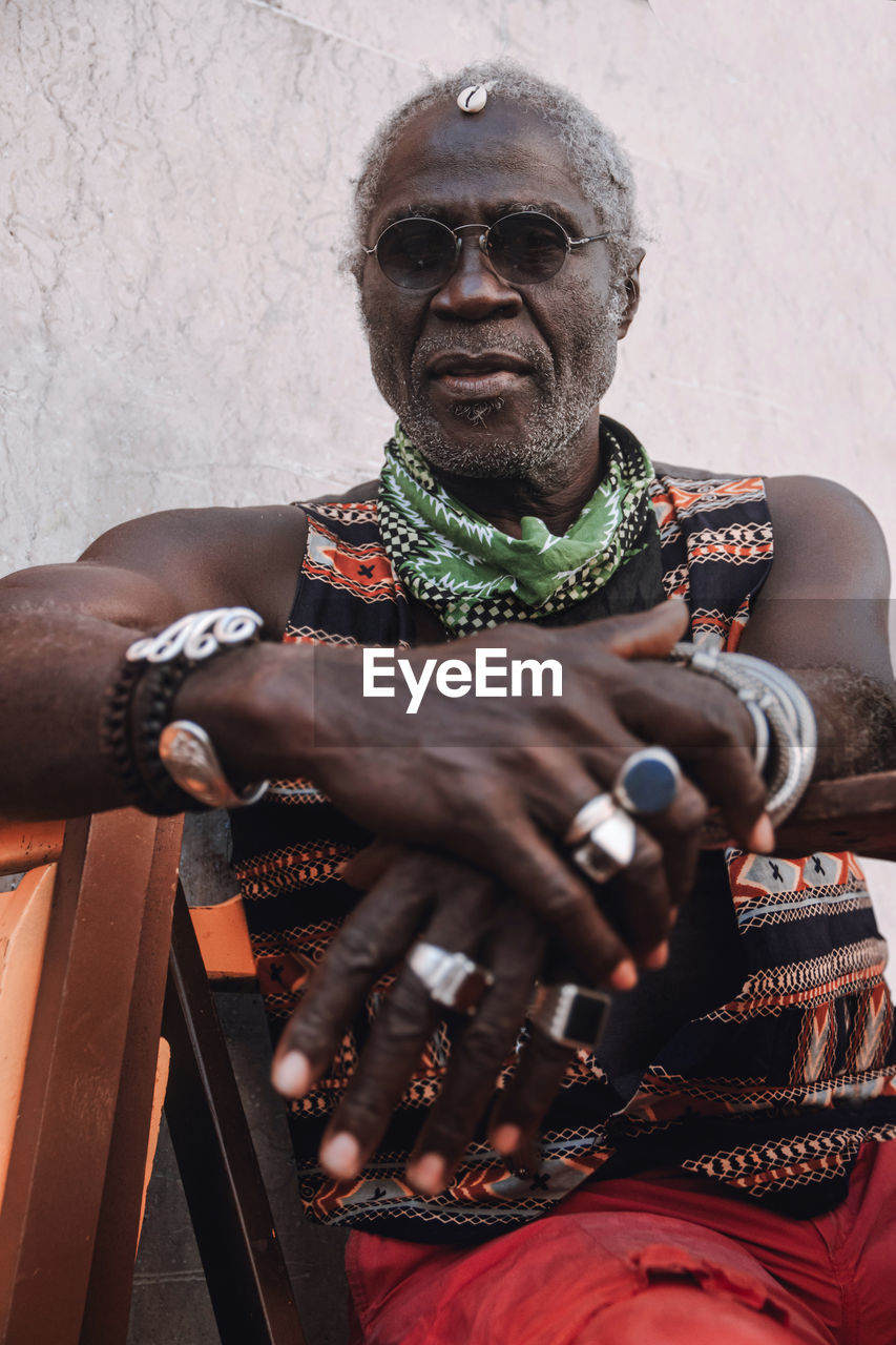 Cool middle aged african american male in rings and modern sunglasses sitting on chair in daylight