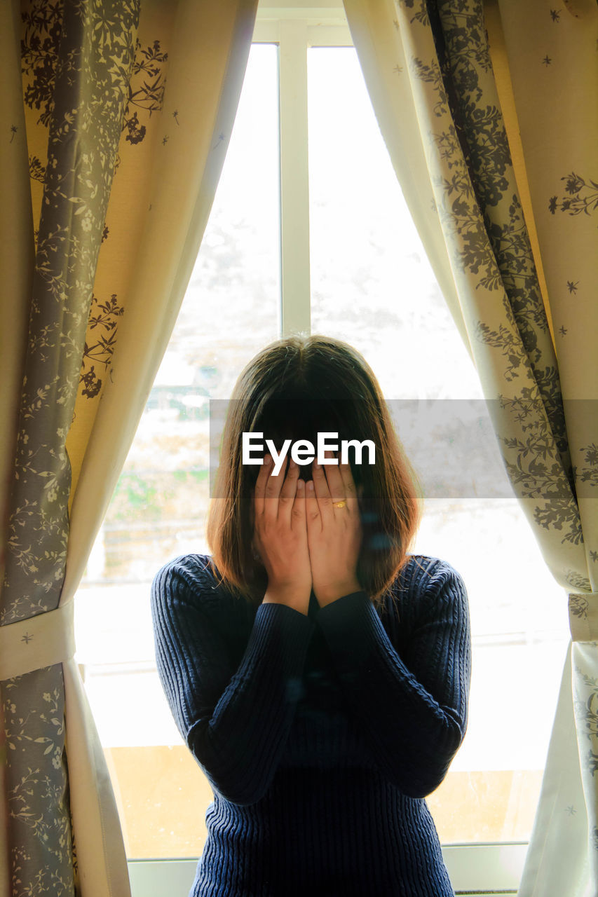 Close-up of beautiful young woman covering face with hands while standing against window