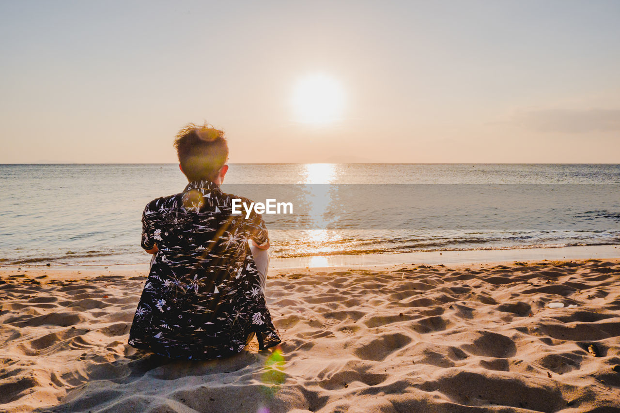 Rear view of man sitting on beach against sky during sunset