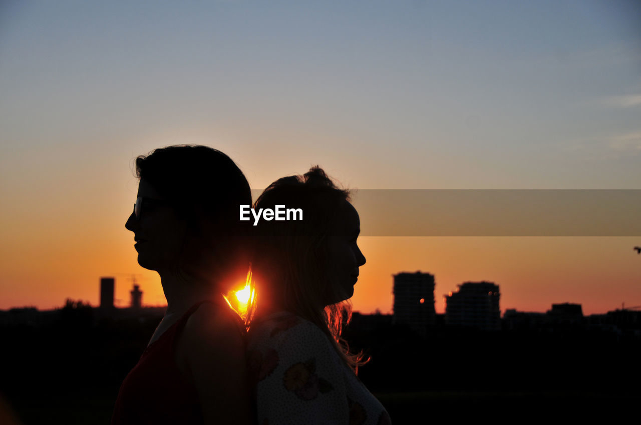 Silhouette of two young women against orange sky in city during sunset. 