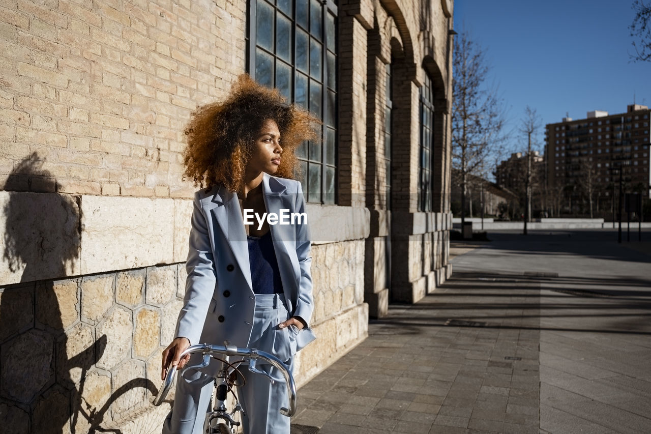 Young woman in blazer standing with bicycle by building during sunny day
