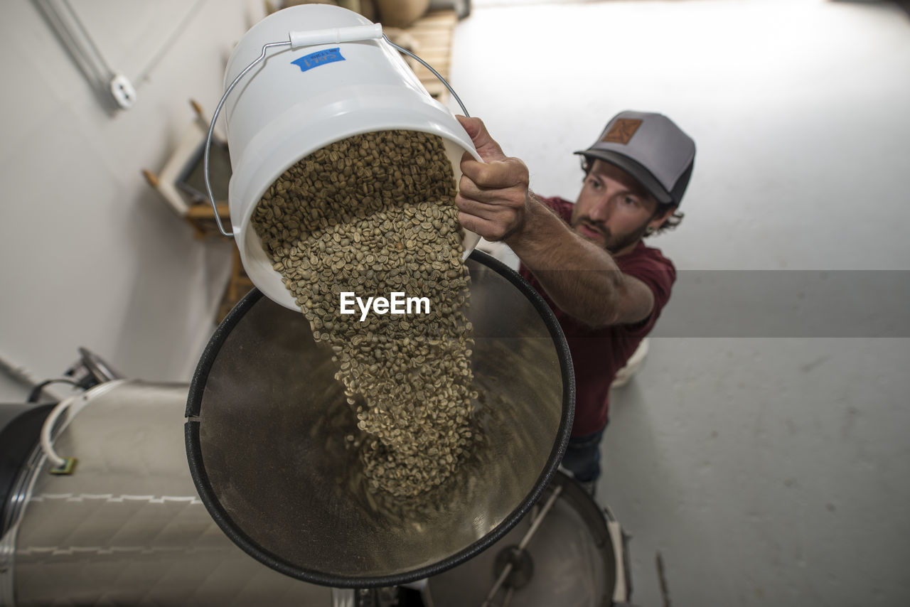 High angle view of man putting raw beans in coffee roaster at shop