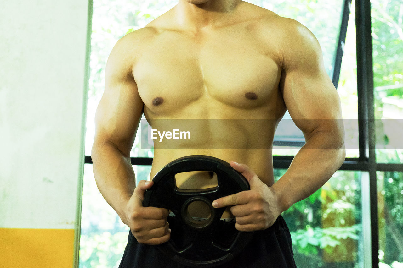 Midsection of shirtless man holding exercise equipment at gym