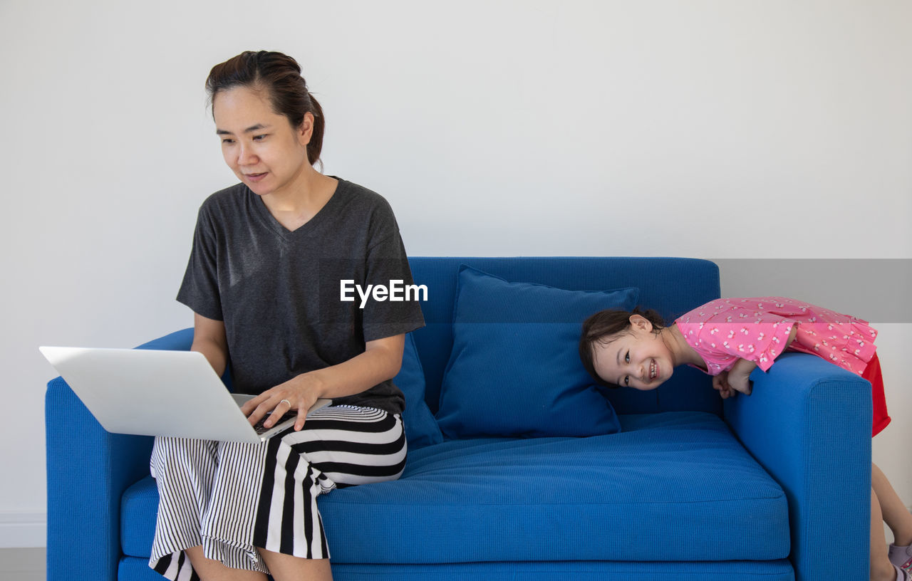Girl playing with mother using laptop on sofa against wall