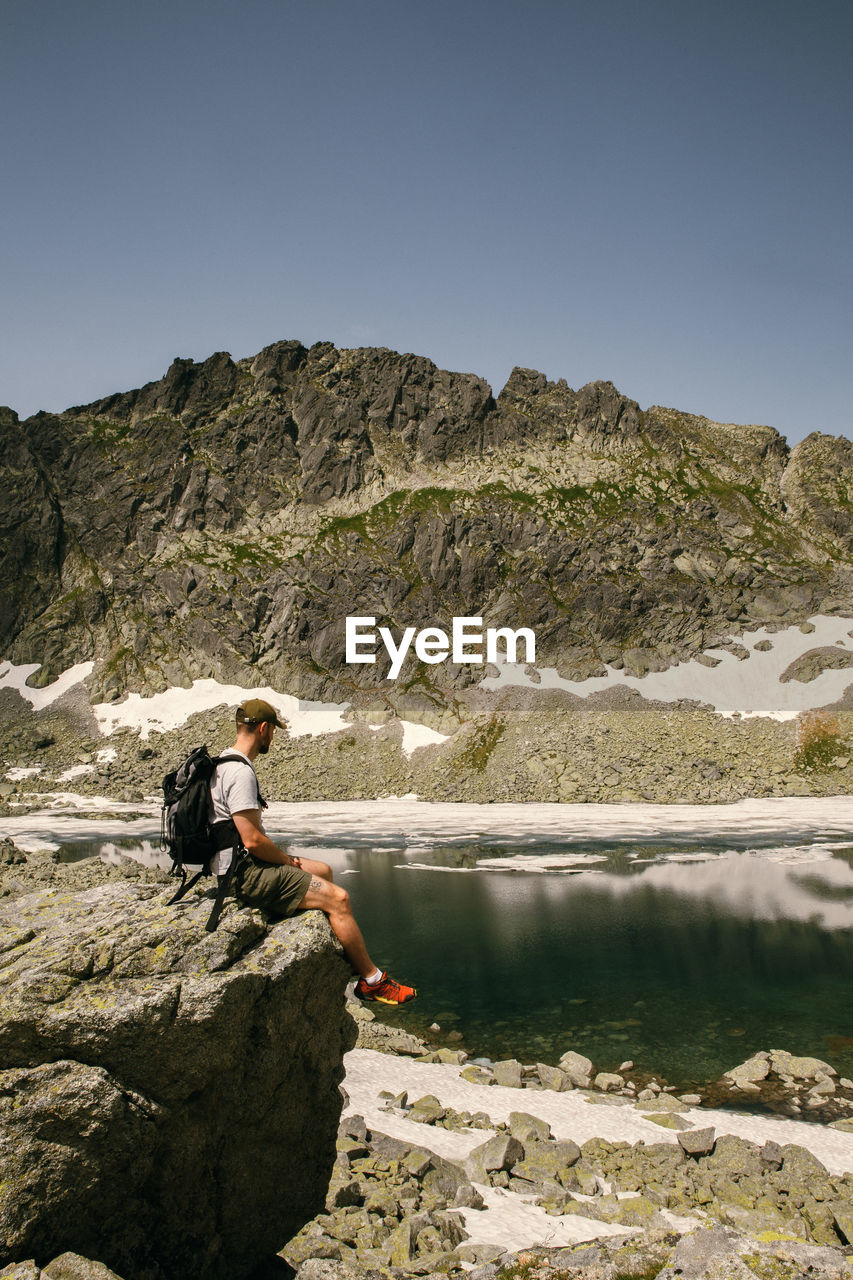 Side view of man on rock by lake against sky