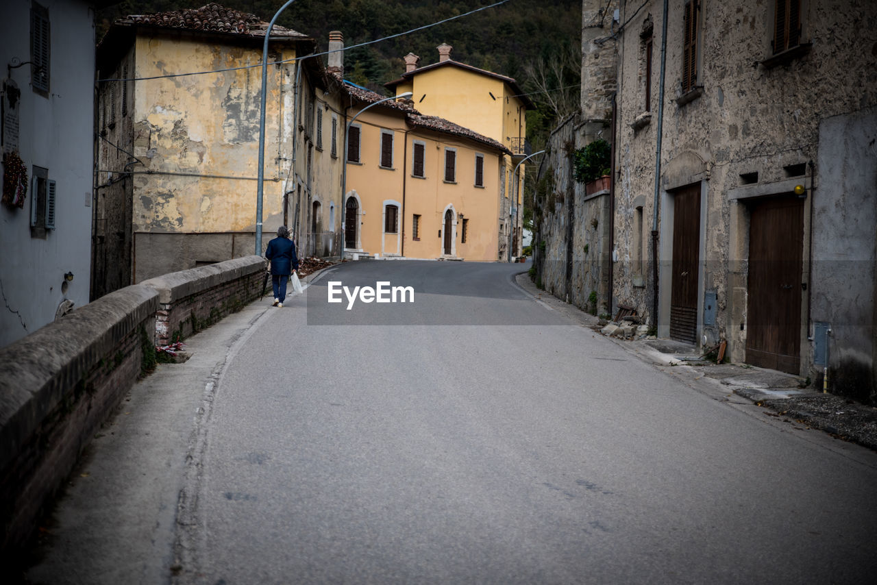 REAR VIEW OF MAN WALKING ON NARROW STREET AMIDST BUILDINGS