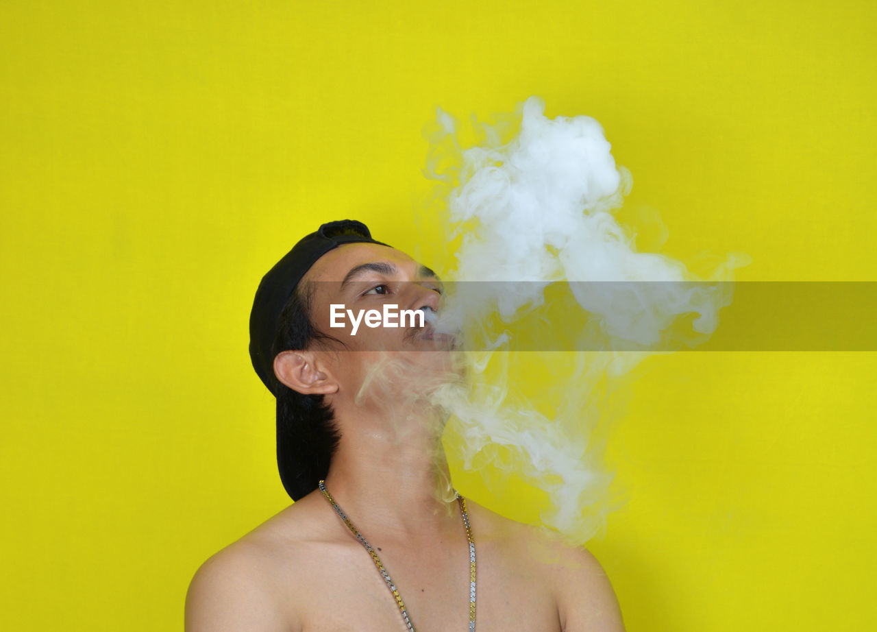 Shirtless young man smoking while standing by yellow background