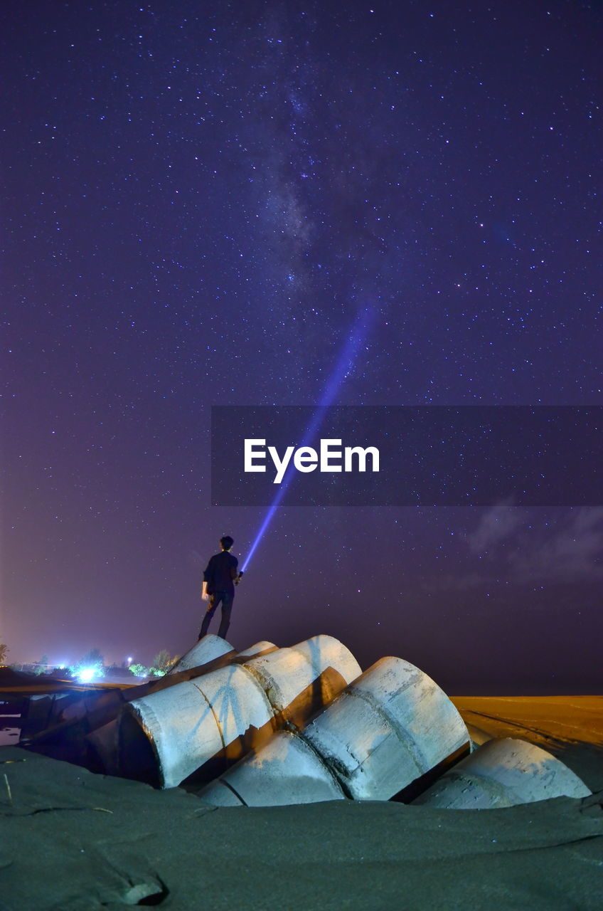 Rear view of man holding illuminated flashlight standing at beach against star field