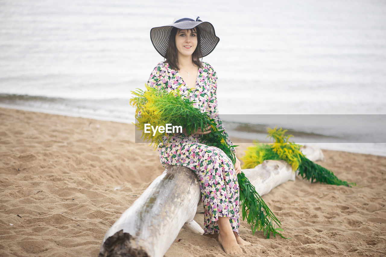 Young woman in summer dress and sun hat sitting on the beach and holding  bouquet of wild flower