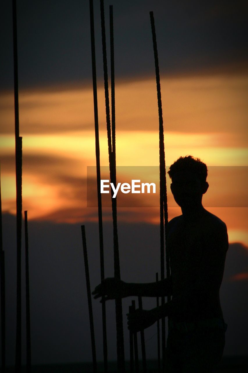 Silhouette man standing by iron rods against sky during sunset